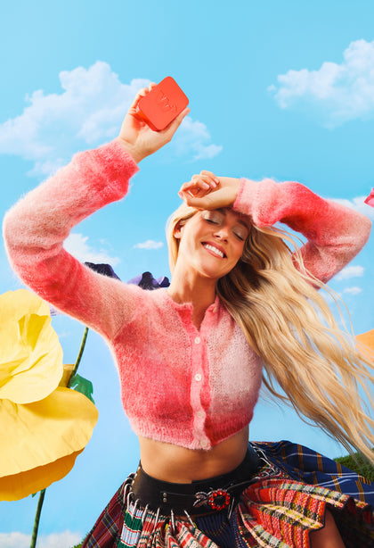 Woman in a pink sweater holding Wizard Oral Strips against a blue sky with clouds and yellow flowers.